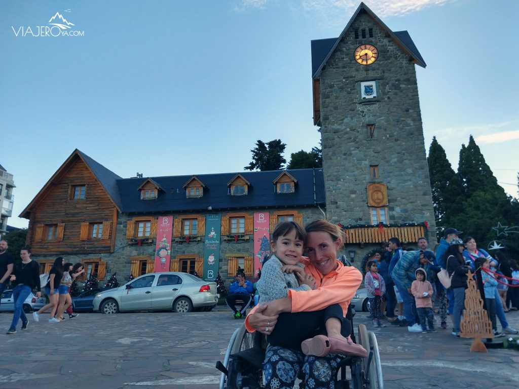 Naty sentada en la silla de ruedas con Sari en su falta y el Centro Civico de Bariloche de fondo